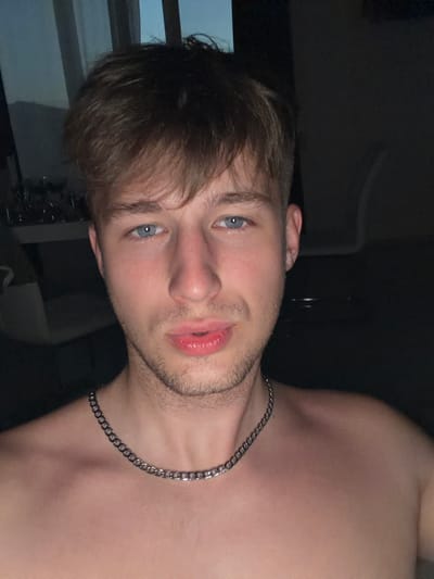 This is a close-up selfie of a young man. He has light brown hair, blue eyes, and fair skin with light stubble. He is wearing a silver chain necklace. The background is dark, suggesting an indoor setting at night or in low light conditions. The environment appears to be residential, possibly an apartment or house, with hints of furniture and windows in the background. The image does not contain any specific visual elements that would indicate a location in the city of Vanino, Russia.