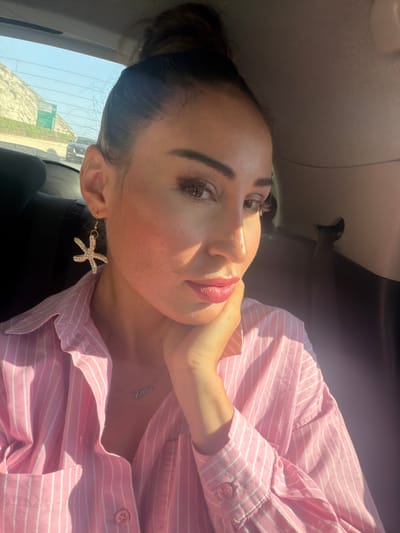 This is a portrait of a woman sitting in a car, with the camera angled downwards. The woman has dark hair pulled back and is wearing a pink and white striped button-down shirt. She is also wearing makeup, including lipstick, eyeshadow, and blush. Her left ear has a starfish-shaped earring with clear gems. A delicate silver necklace with an inscription is visible on her neck.

The lighting suggests it is daytime, with sunlight casting shadows and highlights on her face. The scene appears to be in a vehicle, evidenced by the car's interior visible in the background. Through the rear window, a bright sky and a landscape with some greenery and what appears to be a road sign can be seen, hinting at an outdoor setting.

The woman's pose, with her hand resting on her neck and her head tilted, combined with her makeup, suggests she is posing for the photograph. The overall mood is casual and bright. The location context provided is Aradippou, Cyprus. No visible text is present in the image itself.