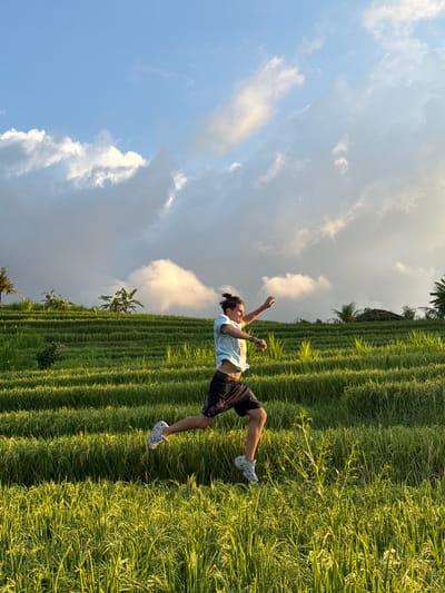 A man in a light blue shirt and black shorts is standing in a lush green rice paddy in Padangbulia, Indonesia. He has his arms raised, seemingly mid-motion. The rice paddy is tiered, creating a stepped landscape. The sky overhead is blue with scattered clouds, some of which are dark.