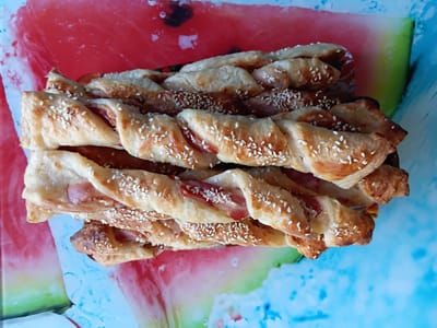 A close-up, overhead shot displays a stack of twisted breadsticks, likely savory, adorned with sesame seeds and pieces of reddish filling, possibly ham or bacon. The breadsticks are golden brown and appear to be baked. They are arranged on a surface with a vibrant, abstract design featuring large swathes of red and green, reminiscent of a stylized watermelon slice, on a predominantly blue background. This surface is likely a placemat or tray. The overall impression is one of a prepared snack or appetizer, presented in a visually appealing manner. There are no people, animals, or any indication of a specific urban environment. The location in Polysayevo, Russia, is not discernible from the visual elements of the image itself.