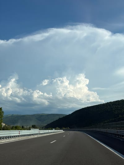 The image shows a highway with a dark asphalt surface and white lane markings. On the left side of the road, there is a metal guardrail with red and white reflectors. Beyond the guardrail, there are trees and hills covered in vegetation. On the right side of the road, there is another metal guardrail, and further in the distance, a dark, densely forested hillside slopes upwards. The sky is a vibrant blue with large, dramatic white cumulus clouds. The road appears to be in a rural or semi-rural setting, possibly on the outskirts of Peshterna, Bulgaria, given the mountainous terrain and vegetation. There are no people or vehicles visible in the immediate scene. The lighting suggests it is daytime, with the sun likely to the right, casting shadows on the hillside. Power lines are visible strung across the landscape.