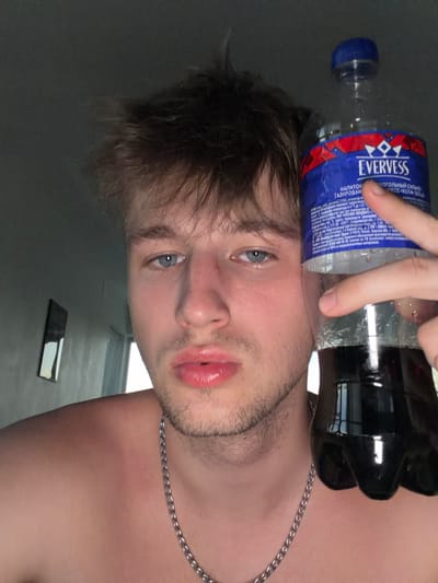 A young adult, possibly male, with light-colored hair and blue eyes, is shown in a close-up shot holding a bottle of carbonated beverage. The person is shirtless and wearing a silver chain necklace. The background is dimly lit with a gray wall and a framed object on it. The beverage bottle has a blue and red label with the brand name "EVERVESS" visible. The image appears to be a selfie taken indoors. There is no specific information indicating the location in Vanino, Russia.