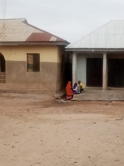 The image depicts an outdoor scene in a residential area, likely in Nigeria given the presence of Nigerian phone number in the prompt. Two buildings, constructed of concrete blocks with corrugated metal roofs, are visible. The building on the left has a tan and brown color scheme, while the one on the right is grey. In front of the grey building, three people are present: two seated and one standing. The person on the left wears an orange hijab, and the person next to them wears a purple hijab. The standing person wears a yellow shirt and black pants. They appear to be interacting with a book or other learning material. The ground is dirt, showing signs of being walked on. The sky is overcast and grey. The image has low resolution.