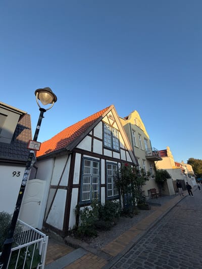 This image captures a narrow cobblestone street lined with historic buildings in Rostock, Germany, likely in the historic city center given the architectural style. The buildings feature traditional German Fachwerk architecture with visible timber framing on a white stucco facade, and red-tiled gabled roofs. One building prominently displays a street number "93" next to a lamppost. The scene is bathed in the warm light of what appears to be late afternoon or early morning sun, casting long shadows across the street and highlighting the textures of the cobblestones and building facades. A few people are visible walking down the street, adding a sense of scale and activity. The sky is clear and blue.