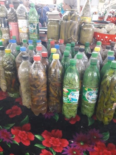 The image displays a vibrant collection of various preserved foods, predominantly contained in repurposed plastic beverage bottles and some glass jars, likely at a market stall or shop. The scene is illuminated by indoor lighting.

Numerous plastic bottles are stacked and arranged, showcasing a variety of contents. Many bottles are filled with green leafy substances, possibly herbs or vegetables, and some of these bottles feature green caps with labels clearly showing the Cyrillic word "КАВЗ" (KAVZ). Other bottles contain green pickled items, resembling gherkins or long beans, while some hold an assortment of pickled peppers in green, red, and orange hues. A few bottles contain a yellowish, more liquid substance. The bottle caps are distinctively colorful, including green, yellow, red, blue, and black. Several larger glass jars on the left also hold preserved items, such as whole pickles. At the top left, a white container with red polka dots holds light-colored, possibly dried, goods.

Partially visible on the far left of the image are at least two individuals, one wearing a light-colored headscarf, appearing to be observing or interacting with the display. The background on the right features a dark fabric adorned with a floral pattern of red and purple flowers with green leaves. The overall arrangement suggests a setting where these preserved items are being sold or presented for consumption, common in local markets or bazaars in regions like Ghafurov, Tajikistan.