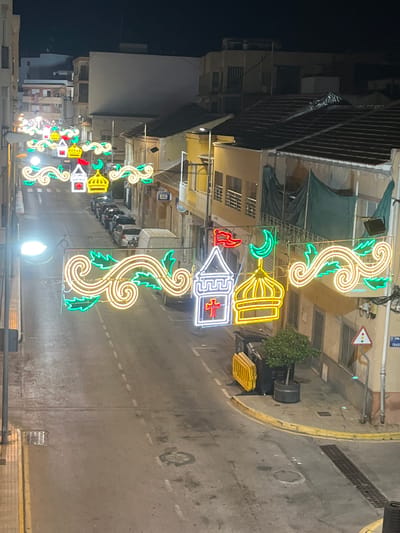 This is a nighttime, high-angle, vertical, medium shot of a street in Rojales, Spain, illuminated by decorative neon lights. The lights are strung across the street and depict festive motifs, including mosque-like structures with crescent moons and crosses. The street itself is wet, suggesting recent rain, and lined with parked cars. Buildings with balconies and awnings border the street, and a street lamp is visible on the left. The overall atmosphere is festive and atmospheric due to the colorful lights against the dark night sky.