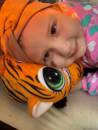 A close-up shot shows a young child lying down with a plush tiger toy. The child has fair skin, brown eyes, and a brown birthmark on her left cheek. She is wearing a light brown headband and a pink jacket with white hearts and green shapes. Her face is partially pressed against the plush tiger, which has orange and black stripes and a large, green and black eye. The background is a beige textured surface, likely a couch or bed. There is no information to discern the location of the scene in the city of Кипчак, Russia, so the location is unknown.