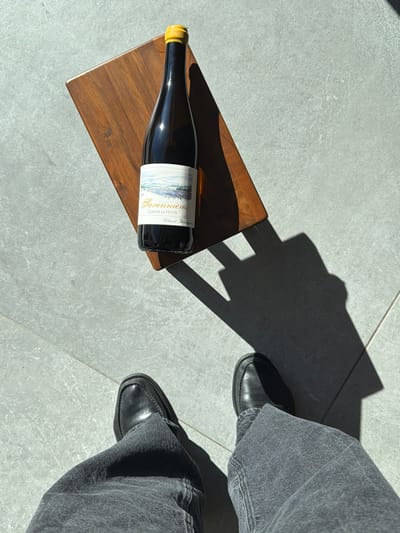 An overhead view in Marche-lez-Écaussinnes, Belgium, depicts the lower legs and feet of an individual wearing dark grey jeans and black leather loafers, standing on a light grey tiled floor. A dark brown wooden serving board is positioned on the floor, angled, with a dark glass wine bottle resting on it. The bottle, sealed with a yellow capsule, features a label identifying it as "Savennières Clos de la Hutte" with a landscape illustration. Strong directional sunlight illuminates the scene, casting distinct, elongated shadows from the board and bottle onto the tiled surface.