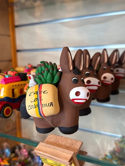 The image displays a clear glass shelf within a retail environment, likely a souvenir shop in the AMCO (Area Metropolitana Centro Occidente) region of Colombia. A primary focus is on a brown ceramic donkey figurine positioned centrally, featuring a white snout with red lips, black eyes, and pointed brown ears with white interior accents. This donkey carries a light beige sack on its back, labeled "CAFE DE COLOMBIA" in black and red text, secured by a string with red, yellow, and blue stripes. A green, spiky plant-like object and a small white chicken figure are placed atop the sack. Behind this lead donkey, a linear formation of identical or similar brown ceramic donkey figurines extends into the background, progressively out of focus, each exhibiting similar facial features. To the left, a portion of a yellow ceramic bus or truck with red wheels and blue details is visible. Below the shelf, a wooden display stand holds a white label partially displaying "PUM COD 154158" and "770313773324". The background comprises additional blurred items on shelves, indicative of merchandise.