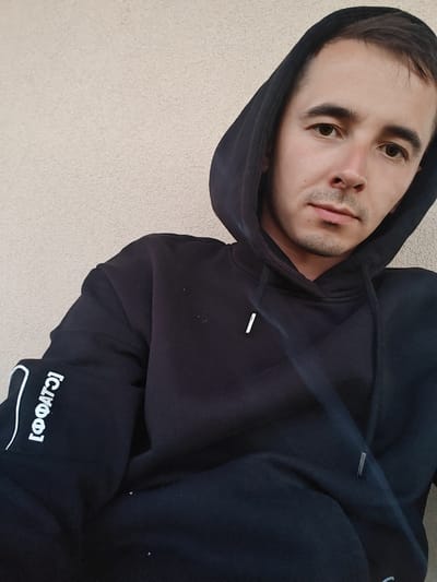 A young man is posing for a close-up selfie, his head and shoulders filling most of the frame. He is wearing a black hooded sweatshirt, with the hood up. The sweatshirt has white text printed vertically on the left sleeve, which appears to be in Cyrillic. The man has short, dark brown hair and brown eyes, with light stubble on his chin. He is looking directly at the camera with a neutral expression. The background is a plain, textured light-colored wall. The image appears to have been taken outdoors during the daytime, as indicated by the natural lighting. The man is positioned slightly to the right of the center of the frame, with the wall occupying the rest of the space. There are no other people or objects visible in the image. The overall impression is casual and personal.