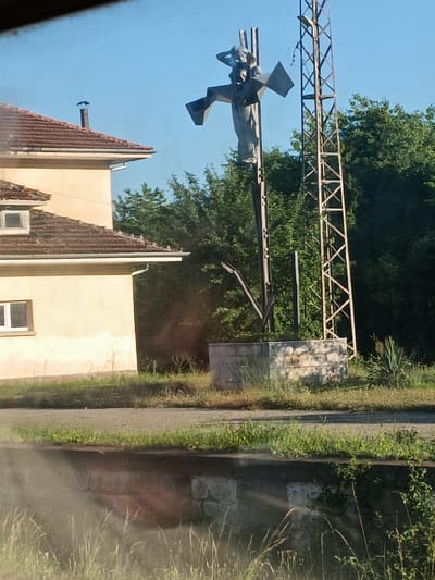 In Bunovo, Bulgaria, a statue of a person is erected on a pole. The statue, made of a gray material, depicts a figure with arms outstretched and head tilted back. The statue is positioned on a metallic pole. A tall, metal structure, resembling a tower, stands to the right of the statue. In the background, lush green trees form a dense canopy. To the left of the statue, a light-colored building with a tiled roof is visible. In the foreground, a stone structure with text is present, partially obscured by vegetation. The ground appears to be a mix of dirt and grass, with some evidence of a paved surface.