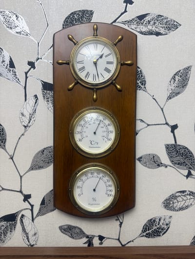 The image shows a wooden plaque mounted on a wall with a patterned wallpaper. The plaque features a clock at the top, designed to resemble a ship's wheel, with Roman numerals and brass-colored hands. Below the clock are two circular instruments. The upper instrument is a thermometer, displaying temperatures in Celsius and Fahrenheit. The lower instrument is a hygrometer, indicating humidity levels as a percentage. All three instruments have brass-colored bezels. The wallpaper has a light background with a repeating pattern of black and gray branches and leaves. The lower edge of the image shows a dark wooden surface, possibly a shelf or a piece of furniture. The scene is indoors and appears to be a home or office setting. There are no people or animals visible in the image. The specific location of Khyrdalan, Azerbaijan, is not visually indicated within the image itself.