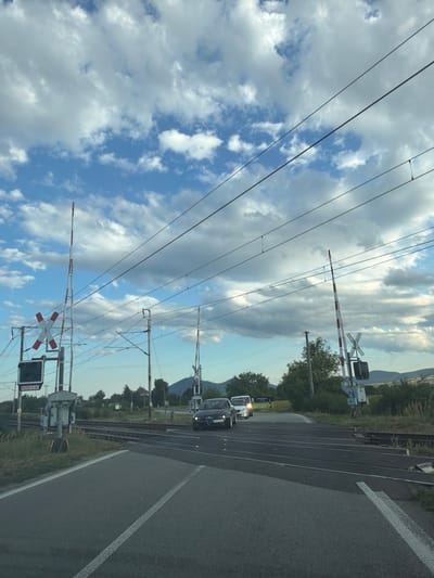 The image depicts a rural road approaching a railway crossing in Čerhov, Slovakia. The sky is filled with dramatic, cloudy formations. Two cars are visible on the road: a dark-colored sedan on the right and a white van further ahead and to the left. The railway crossing features multiple barriers, traffic lights, and overhead electrical wires. The landscape is characterized by grassy fields, trees, and distant hills. The scene appears to be captured during the late afternoon or early evening.