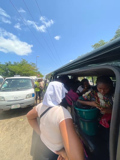 The image shows a street scene in Gbongan, Nigeria, on a sunny day. In the foreground, the back of a person wearing a light-colored t-shirt and a white towel draped over their head is visible. This person is facing away from the camera and appears to be boarding or exiting a vehicle.
To the left of the foreground subject, a white minivan is parked. The minivan has a license plate that reads "LES 31XC" and the front grille has a Mazda logo. The minivan's doors are open, and several people are visible inside and around it. A man in a bright green and yellow patterned shirt and dark shorts is standing next to the minivan, looking at his phone. Another man, wearing a striped shirt, is standing behind him.
To the right of the foreground, the interior of a dark-colored passenger van is visible. Two women are seated inside. One woman, wearing a colorful patterned top, is holding a smartphone and looking at it. Next to her, another woman is eating something and looking forward. A green bucket is placed on the floor of the van. Several bags are visible inside the van, including a pink and black patterned bag.
The background of the image shows trees and other vehicles. Power lines are visible against a blue sky with scattered white clouds. The overall scene appears to be a busy street or bus stop area.