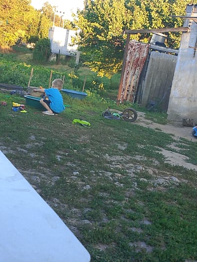 The image depicts a backyard scene in Pryvilne, Ukraine, on a sunny day. A young boy, wearing a blue t-shirt and dark shorts, is crouched in a green plastic tub filled with water. He is facing away from the camera, towards the left side of the frame, and appears to be playing with toys. Scattered around the tub are several colorful toys, including a yellow truck and a blue car. In the background, there is lush green vegetation and trees, suggesting a rural or semi-rural setting. A dilapidated structure, possibly a shed or outbuilding, is visible on the right side of the image. The ground is covered in grass and dirt. A toy scooter with a large wheel is lying on its side in the middle of the yard.