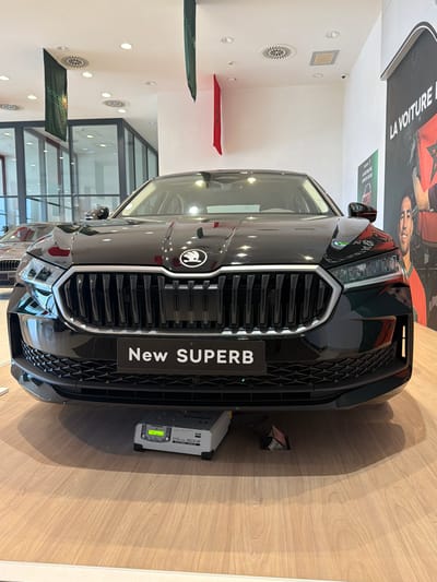 The image depicts the front of a black Skoda "New Superb" car displayed in a showroom. The car faces the viewer with its grille prominently featuring the car's name. Next to the car sits a GYS FLASH 100.12 HF device, possibly a battery charger or diagnostic tool. The background features the interior of the showroom with visible flags and lighting. A sign in the background mentions "La Voiture" and "SICDA", with additional text in French. The specific location is the city of Ouahat Sidi Brahim, Morocco.