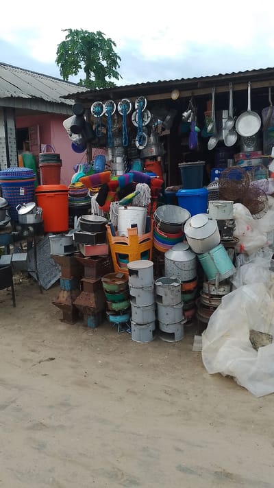 This is a photograph of an outdoor market stall in Agbarho, Nigeria. The stall is filled with a variety of kitchenware and household items, including pots, pans, buckets, basins, and brooms. The items are stacked high and arranged in a haphazard manner, suggesting a busy and active marketplace. The stall appears to be made of corrugated metal and wood. In the background, a pink building and a large tree are visible. The ground is dusty and uneven, typical of a Nigerian street. There are no people visible in the image. The overall impression is one of a vibrant and bustling local market.