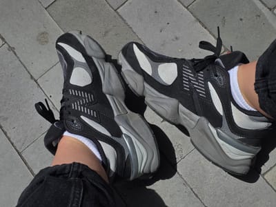A pair of black and grey New Balance 9060 sneakers are shown from a top-down perspective. The sneakers are worn by an unseen individual with white socks and dark denim pants. The sneakers are positioned on a grey tiled surface, characteristic of an outdoor urban environment, possibly a promenade or plaza. The lighting suggests it is daytime, with distinct shadows cast by the shoes. The tiled ground has some minor marks and stains, indicative of foot traffic. The overall scene is static, focusing on the footwear against the urban backdrop. The location in Puerto De La Cruz, Spain, is not explicitly indicated by any visual cues within the image itself.