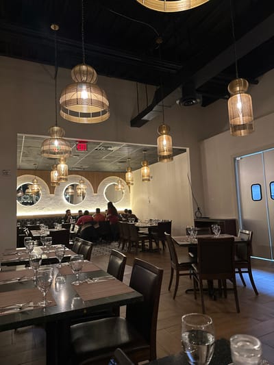 A restaurant scene shows tables set with glasses and cutlery, ready for diners. Some people are seated. Above, decorative globe-shaped lights hang on chains. Mirrors adorn a wall, reflecting the space. An "EXIT" sign is visible. The room has dark-toned furniture and warm lighting, creating an inviting atmosphere. The perspective is tilted, offering an unusual view of the restaurant interior.