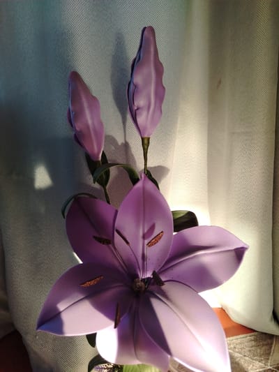 A close-up, high-angle shot shows a purple, artificial lily with three buds and one open bloom. The flower is positioned in front of a light-colored, textured curtain. The lighting creates strong shadows and highlights on the petals and the curtain. The scene is indoors, likely a domestic setting, and there is no indication of the location being Zyryanskoye, Russia.