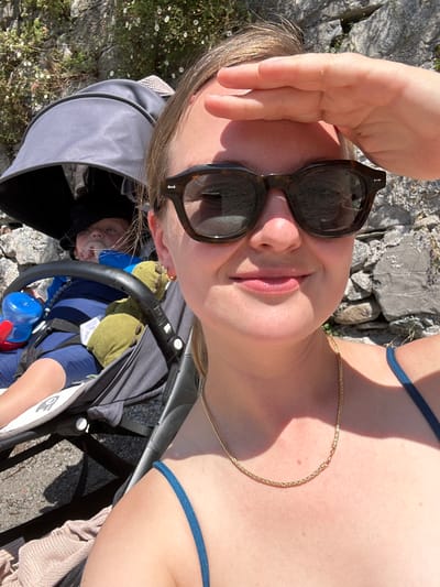 A selfie-style photograph captures an adult female and an infant in a stroller, situated outdoors in Abbadia Lariana, Italy. The female, positioned in the foreground, wears dark, tortoise-shell sunglasses and a gold-colored chain necklace over a dark blue-toned garment strap. Her right hand is raised to shield her eyes from the bright sunlight. Her expression displays a subtle smile. To her left and slightly behind her, an infant is seated in a gray stroller equipped with a dark canopy. The infant wears a dark head covering, and a blue sippy cup with a red spout is visible on the stroller's left side. A green stuffed animal rests next to the infant. The background consists of a sunlit rock face with sparse vegetation, including small white flowers. The ground visible beneath the stroller is dark, partially covered by a light-colored textile.