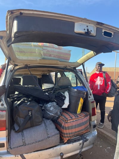 The open trunk of a silver car is packed with luggage, including duffel bags and a checkered suitcase. A black bucket sits atop some items. A man in a red skeleton sweater stands nearby with his back to the car, while another person stands partially visible on the right side of the frame. The car appears old and slightly worn, and the scene is set outdoors during daylight.