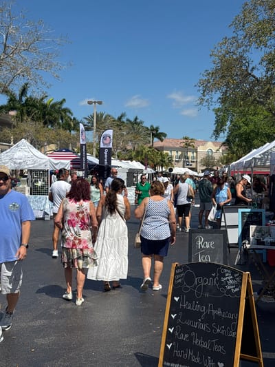 An outdoor market is depicted under a clear blue sky in Pelican Marsh, United States. Numerous individuals, predominantly adults, are present, moving along a paved thoroughfare lined with white pop-up canopy tents serving as vendor stalls. The environment is well-lit by direct sunlight.

In the immediate foreground, a black A-frame chalkboard sign stands on the right, advertising "Rare Tea" and "Organic" products including "Healing Apothecary," "Luxurious Skin Care," "Rare Herbal Teas," "Hand Made With," "New Tallow Soap," and "New Tallow Butter." Several people are visible in various stages of walking. A man in a blue t-shirt and light shorts is partially visible on the far left, facing the camera. Three women are prominent in the central foreground, seen from behind: one in a floral patterned dress, one in a white maxi dress, and one in a blue patterned top and dark shorts.

Further down the market pathway, more people are gathered around the stalls. Two tall, vertical flags displaying "Operation Vet Help" with an American flag motif are visible behind the tents on the left side. Another American flag banner is partially seen. In the background, low-rise buildings with light-colored walls and tiled roofs are partially obscured by lush green and some bare-branched trees, including numerous palm trees. Streetlights are also visible above the tree line.