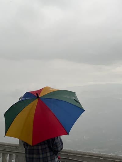 A view of a cloudy, overcast sky and the edge of a mountain, captured from the city of Rathmalkaduwa, Sri Lanka. A multi-colored umbrella, predominantly blue, red, yellow, and green, is prominently featured in the frame, held by an unseen person wearing a plaid shirt. A railing or low wall is visible on the right edge of the frame.