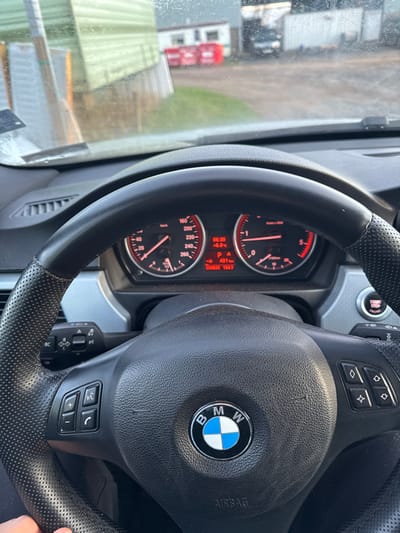 The scene is viewed from inside a BMW vehicle, looking through a smudged windshield at an outdoor environment in Inchture, United Kingdom. A black leather multi-function steering wheel with the BMW logo is prominent in the foreground, with a hand holding its lower-left perforated section. The instrument cluster, illuminated in red, displays 06:30 AM, an external temperature of +6.0 °C, a remaining range of 401 km, and an odometer reading of 290,893 km. Outside the vehicle, a dirt and grass-covered yard extends into the mid-ground. A large, light green corrugated metal building is visible on the left side of the view. In the background, a row of red skip bins is present, situated in front of white industrial structures. A dark-colored vehicle is partially visible further back, near these white buildings.