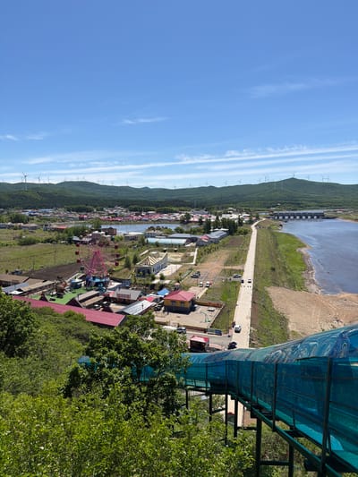 This elevated view captures a vibrant town or village scene in Jiejinkou, China, nestled between rolling green hills and a body of water under a clear blue sky. The image is taken from a high vantage point, looking down over the landscape.

In the background, a series of lush, forested hills stretch horizontally across the left side of the frame, adorned with numerous white wind turbines along their ridge, signifying renewable energy generation. To the right of the hills, a long, narrow body of water, possibly a river or reservoir, is visible. A dark, linear structure, possibly a dam or bridge, spans across the water, separating it from a wider, drier bank further to the right.

The mid-ground is dominated by the town, featuring an array of colorful buildings with various roof types. A paved road snakes through the settlement, running parallel to the hills and the water. Several cars are visible on and beside this road. A prominent pink ferris wheel stands out in the center-left of the village area, suggesting a recreational or amusement zone, with other smaller rides or structures nearby. Scattered trees and green spaces are interspersed among the buildings.

In the immediate foreground on the right, part of a green metal structure with a translucent blue corrugated roof is visible, suggesting a covered walkway or viewing platform from which the photo was taken. This structure is partially framed by dense green foliage from nearby trees.

The scene depicts a bright, sunny daytime, indicated by the clear blue sky with a few wispy, streaky clouds. There are no specific activities or events prominently depicted, but the presence of the ferris wheel and the overall setup suggests a lively, possibly tourist-oriented area. No discernible text is visible in the image.