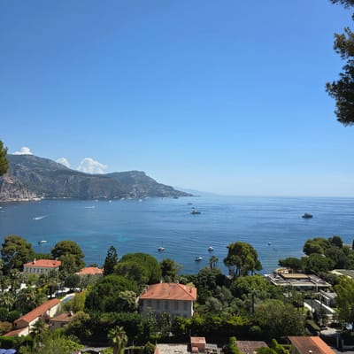 This is a high-angle, daylight, medium shot of a coastal view in Saint-Jean-Cap-Ferrat, France. The scene is dominated by a vibrant blue sea and a clear, bright blue sky with a few scattered white clouds. In the foreground, a lush green landscape with various trees and foliage is visible, interspersed with several large, modern, and traditional houses with terracotta roofs. Some of these residences appear to be villas, complete with swimming pools and manicured gardens. Several boats, including sailboats and yachts, are scattered across the calm blue water. In the background, a prominent, steep, mountainous coastline rises from the sea, dotted with small structures and vegetation. The overall impression is of a luxurious and scenic Mediterranean setting.