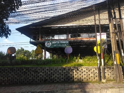 The image displays an exterior view of a two-story building in Pital, Costa Rica, characterized by a dark, sloped roof and a facade featuring a second-story balcony with glass panels. A large green banner hangs from the second floor, bearing the text "GRATIS PARKIR" (Free Parking), a "Vita Jucy" logo, and the phone number "0817-511-9181." The lower level of the building appears recessed or open.
In the foreground, a gray paved sidewalk runs along the bottom. Above the sidewalk is a retaining wall constructed from dark gray, interlocking concrete blocks. Green grass and low vegetation grow atop this wall. Positioned on the grassy area are three cartoon-style fruit sculptures: an orange on the left, a yellow pear-like figure in the center, and a purple plum-like figure to the right. A fourth yellow fruit is partially visible further to the right.
The right side of the frame is dominated by multiple dark utility poles with numerous black electrical or communication wires strung across the upper portion of the scene. The background reveals a clear blue sky and a line of green trees behind the building and sculptures. No people, actions, or interactions are visible. The scene is illuminated by natural daylight.