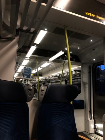 The interior of a train is shown. Seats with blue patterned fabric face forward with a black headrest. A yellow pole runs horizontally between the seats and the window. Overhead lighting illuminates the interior. Outside the window, a sign reads "JOCHFELDEN". The next stop is displayed on a digital display at the front of the train. A information board is on the wall.