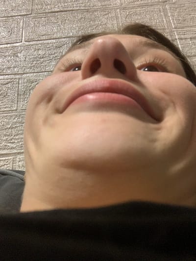The image is a close-up, low-angle shot of a person's face, specifically focusing on their nose, mouth, and eye. The person is wearing dark clothing, partially visible on the left side of the frame. The background appears to be a textured wall with a repeating rectangular pattern, possibly wallpaper or decorative tiles. The lighting is soft, and there are no clear indications of the time of day or weather. The person's expression is difficult to discern due to the extreme close-up and angled perspective, but their mouth is slightly open. There is no visible text in the image. The location context is Belogorsk, Russia.