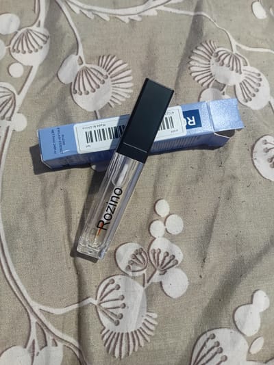 A clear glass tube with a black cap and a white applicator wand is positioned diagonally across a light-colored fabric with a white floral pattern. The tube has the brand name "Rozino" printed in black lettering. To the upper right of the tube, a small blue box with white lettering is partially visible. The box has text that includes "Made in China" and "RO" in large blue letters. The scene is lit by sunlight, creating shadows. The location is not identifiable as Bocani, Moldova.