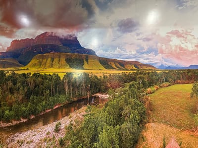 The image depicts a panoramic view of a dramatic mountain landscape. In the background, imposing, sheer-faced mountains rise, some shrouded in clouds. The slopes leading up to these peaks are covered in lush green vegetation, transitioning to golden grasslands on lower terraces. A dense forest occupies the midground, with a winding river cutting through it. The riverbed is composed of smooth, light-colored stones. The sky above is filled with a mix of dramatic clouds, some tinged with pink and orange from the setting sun, and others a more muted gray. The overall impression is one of a wild and remote natural environment. There are no people or structures that are indicative of the city of Torrejón de Ardoz, Spain, or any urban environment present in this natural landscape.