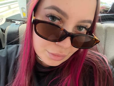 A close-up selfie of a young woman with bright pink hair, wearing sunglasses, a nose ring, and a pearl necklace. She is seated in what appears to be a vehicle, indicated by the seatbelt buckle and textured seat visible in the background. The image is focused on her face and upper body, with her hair framing her face. The lighting suggests an indoor or shaded outdoor environment. The location of Teshebs, Russia, is not discernible from the visual information in the image.