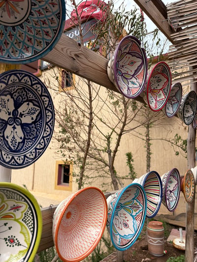 The image depicts an outdoor display of Moroccan pottery in Zerkten, Morocco. The pottery, a mix of bowls and plates, is arranged on a wooden structure. The items showcase traditional Moroccan designs with vibrant colors. In the background, a light-colored building with a window is visible, partially obscured by tree branches. Other elements include a reed roof structure.