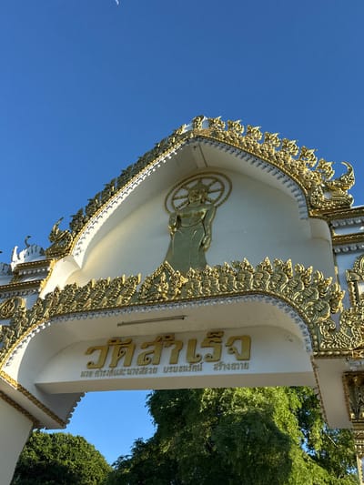 The image shows a close-up view of a white and gold temple archway with intricate decorative carvings. A golden statue of a standing Buddha with a halo is visible in the upper center of the arch. Below the Buddha, golden Thai script is inscribed. The archway is adorned with elaborate gold ornamentation featuring mythical creatures and floral motifs. The background consists of a clear blue sky with a crescent moon faintly visible in the upper left corner. Lush green trees are seen through the opening of the archway. The scene appears to be a temple entrance in Baan Tha Po, Thailand, suggesting a cultural or religious site.