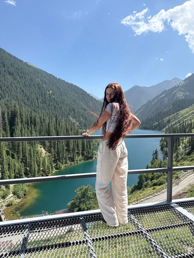A young woman with long, dark brown hair poses for a photo on a metal walkway overlooking a stunning mountain lake in Kurmeti, Kazakhstan. She is wearing a light-colored, short-sleeved crop top and loose-fitting cream-colored pants. Her arm is bent, with her hand resting on her hip. The walkway has a metal railing and a mesh metal floor, allowing a clear view of the breathtaking scenery below and around.
The lake, a vibrant turquoise blue, is surrounded by steep, densely forested mountains. The trees, predominantly evergreens, blanket the slopes, creating a rich, textured landscape. In the distance, larger, more rugged mountains rise, their peaks possibly shrouded in a light haze. The sky is a clear, bright blue with a few wispy white clouds, suggesting a pleasant, sunny day. The time of day appears to be late morning or early afternoon, given the brightness and the angle of the shadows. The woman's pose and facial expression suggest confidence and enjoyment of the magnificent natural setting. The presence of the walkway indicates a developed viewpoint or possibly a bridge, providing access to this elevated perspective of the valley.