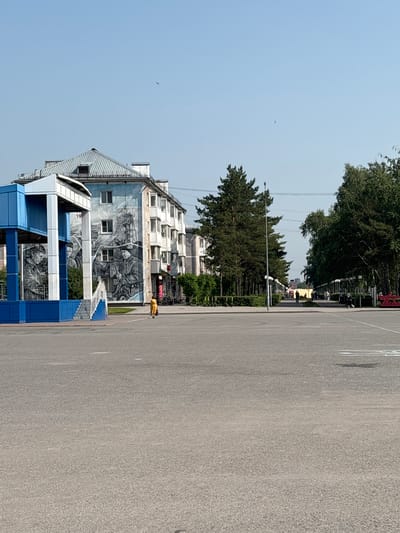 A wide, empty asphalt plaza occupies the foreground of the image, with faint white markings visible on its surface. To the left, a bright blue and white structure with a covered roof and open sides stands prominently. This structure features a mural on its side, depicting several figures in black and white. A set of stairs leads up to this structure. Adjacent to the blue and white structure is a multi-story building with a mural covering its facade. The mural is in black and white and depicts scenes of industry or labor, with figures working. The building itself has white-framed windows and balconies on its upper floors. A single person in a yellow outfit is walking on the asphalt in front of the mural.

Beyond the immediate area, a row of tall, dark green pine trees lines a pathway. Further down this pathway, several people are walking, and on the right side of the pathway, a line of trees with lighter green foliage creates a dense barrier. The sky above is clear and light blue, with a few distant birds visible. The scene appears to be an urban outdoor space, likely a public square or park area in the city of Belovo, Russia, given the architectural style and the mural's theme, which might represent the city's history or local heritage.