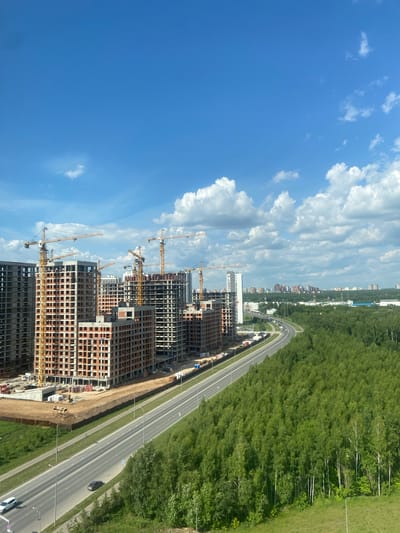 The image depicts a construction site in Kazan, Russia, on a clear day with scattered clouds. Multiple high-rise buildings are under construction, characterized by exposed brickwork and visible scaffolding. Numerous yellow tower cranes are actively engaged in the construction process. A multi-lane highway runs alongside the construction site, with a few vehicles visible on it. Adjacent to the highway and construction zone is a dense forest of green trees, with some trees exhibiting white bark, likely birch. In the distance, a city skyline with more buildings can be observed, suggesting an urban or suburban setting. The overall scene is one of ongoing development within a semi-natural environment.