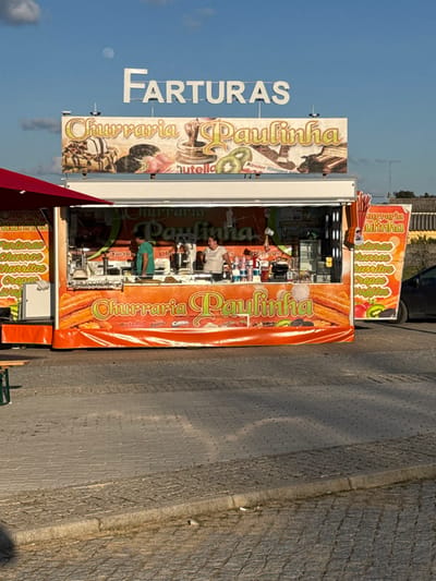 The image displays a "Churraria Paulinha" food truck situated in Mora, Portugal. Above the white food truck, large white letters spell "FARTURAS" against a clear blue sky containing a visible moon and scattered white clouds. The truck's exterior is extensively branded with orange and yellow signage, featuring "Churraria Paulinha" text and images of churros, waffles, Nutella, strawberries, kiwi, Oreo cookies, and Kinder Bueno products. Inside the open serving area, a male in a green t-shirt is positioned on the left, and a female in a white t-shirt is on the right, both standing behind the counter, appearing engaged with food preparation equipment. To the left of the truck, a red umbrella awning extends over a partially visible green wooden picnic table. The ground in the foreground is paved with light grey paver stones, exhibiting prominent shadows indicative of a low sun angle. A raised curb separates sections of the paved area. A dark-colored car is partially visible behind the truck on the right, with a light-colored building in the distant background.