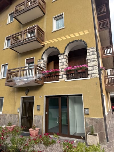 The image shows the exterior of a multi-story building in Cavedago, Italy, with a mustard-yellow stucco facade. The building features several wooden balconies with railings, some of which are adorned with planters filled with pink flowers. Two arched openings with stone accents are visible on the second floor, also decorated with planters. A dark brown door is situated on the ground floor, next to a glass-paneled entrance. Potted plants and flowering shrubs are arranged in front of the building. The scene is captured from a slightly upward angle, with the building's corner prominently displayed.