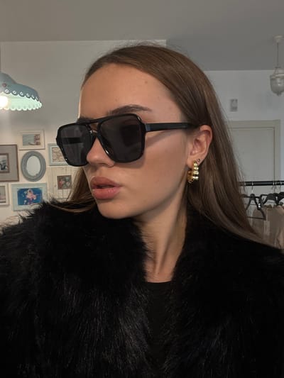 An eye-level indoor photograph, captured in Kfar Yona, Israel, depicts a female subject from the chest up, angled to her left. She possesses long brown hair and wears black-framed, dark-lensed sunglasses featuring a double bridge design. A multi-ridged gold-toned hoop earring is visible on her right earlobe. She is dressed in a black faux fur jacket. Her lips are slightly parted. The background consists of a plain white wall. To the upper left, an illuminated white scalloped light fixture with a floral motif is visible. Below it, a varied arrangement of framed wall decor, including rectangular pictures with light-toned scenes and a circular mirror, is present. To the upper right, a white ceiling light fixture is partially visible, alongside a metal clothing rack displaying several light-colored garments with visible tags.