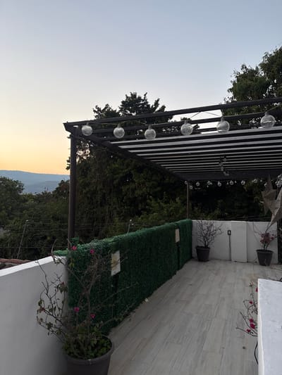 An outdoor patio features a black pergola adorned with string lights, set against a twilight sky and mountainous horizon. A green faux hedge line provides privacy, alongside white walls accented with potted plants. The patio floor is tiled in light grey, enhancing the space's clean lines. The scene presents a tranquil ambiance, ideal for relaxation.