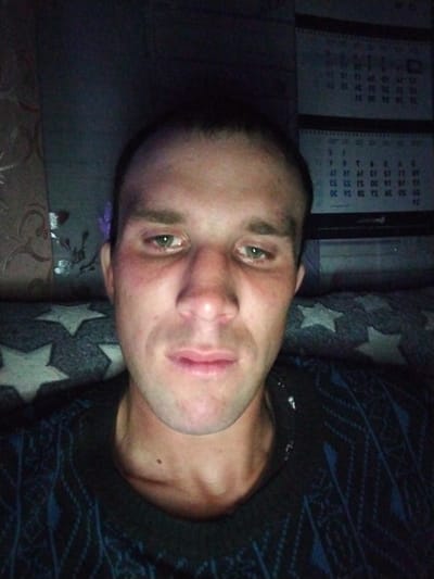 This is a dimly lit, indoor, eye-level, waist-up selfie of a young to middle-aged Caucasian male with light skin and short, dark hair. The man is looking directly at the camera with a neutral expression. His eyes are green, and he has prominent cheekbones and a slightly crooked nose. He is wearing a dark blue sweater with a geometric pattern.

In the background, on the left, there is a patterned curtain or fabric with a floral design. To the right of the man, there are two calendars hanging on the wall, displaying dates and numbers. The overall environment appears to be a room with low lighting, likely a residential setting in the village of Zharabkovichy, Zharabkovichy Rural Council, Belarus. The lighting conditions make it difficult to discern finer details of the environment.