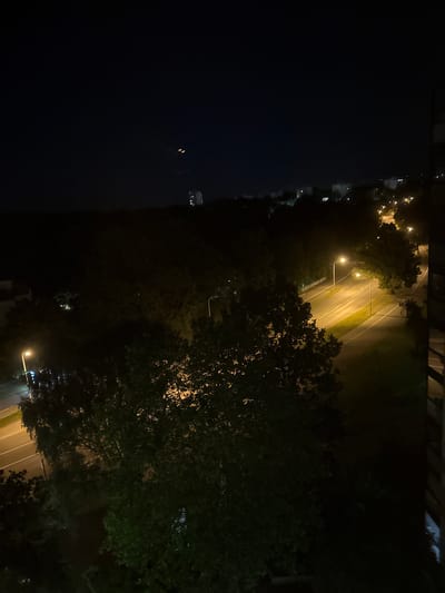 The image depicts a nighttime urban scene in Wolfsburg, Germany, viewed from an elevated position, likely a window. In the foreground, dense foliage of trees is visible, partially obscuring the view. Beyond the trees, a well-lit street runs through the scene, with streetlights illuminating the asphalt and white lane markings. The street appears to be lined with grass and more trees. In the distance, several buildings are visible, some with illuminated windows, indicating residential or commercial structures. The sky is dark, suggesting late evening or early morning hours. Two small, bright lights are visible in the upper portion of the sky, potentially aircraft or drones.