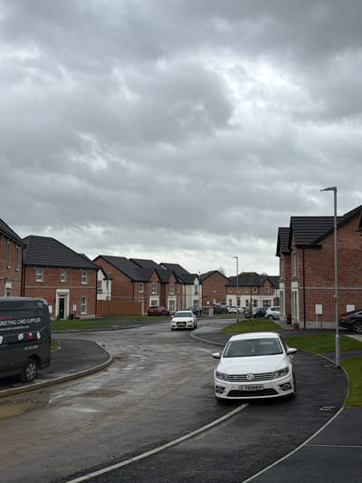 The image shows a residential street in Craigavon, United Kingdom. The scene depicts a row of two-story brick houses with dark grey roofs under an overcast sky. The street is wet, indicating recent rain. Several vehicles are visible, including a white SUV with license plate "CT60NER" parked on the right side of the road, a white car further up the street, and a black van with "Greeting Card Supplier" and website information on the side parked at the front. Streetlights are positioned along the roadside. There are no people visible in the image.