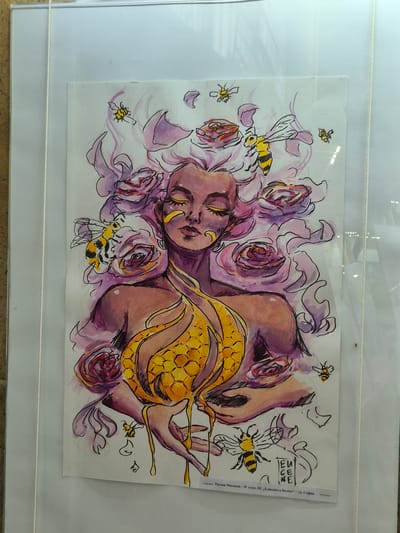 The image depicts a watercolor drawing of a woman with a honeycomb draped over her torso. Her head is surrounded by purple roses, and several cartoon-like bees are scattered around her. The drawing has a signature or logo on the top right and some text along the right edge, visible in the top right corner. The drawing is mounted in a frame, likely a display. The image does not provide any information regarding its location in Srebarna, Bulgaria.